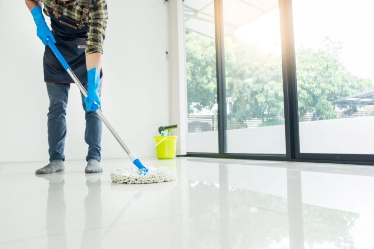 Young housekeeper cleaning floor mobbing holding mop and plastic bucket with brushes, gloves and detergents in the leaving room house floor helping his wife